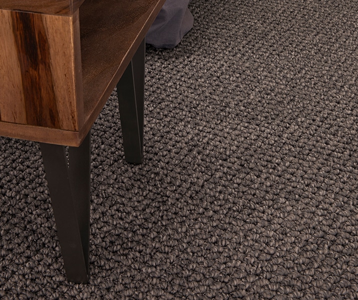 A close-up of Reside Green Cape Montebella polyester carpet flooring in a bedroom setting. The carpet features a rich, textured loop pile in a deep charcoal tone, offering a modern and sophisticated look. The flooring is paired with a timber bedside table, adding a natural contrast that enhances the cosy and contemporary design, ideal for Australian interiors prioritising durability and style. Modern living room with textured carpet – Flooring Xtra
